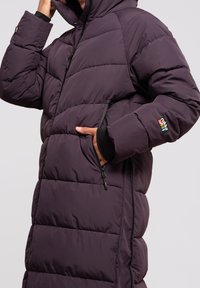 Lilla pufferjakke med hætte, lynlås foran og sidelommer. Materialet er quiltet med en glat overflade og kontrasterende sorte detaljer.