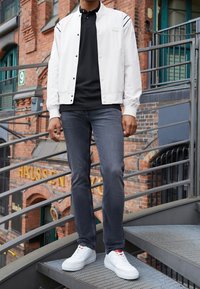 Veste bomber blanche par-dessus un polo noir, jeans en denim foncé et baskets blanches avec des accents rouges, se tenant sur des escaliers métalliques dans un environnement urbain.