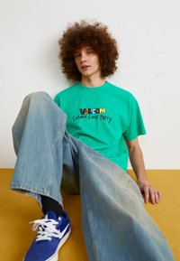Turquoise cotton t-shirt with "Volcom Cosmic Love Party" graphic, paired with light blue wide-leg jeans and blue sneakers. Sitting pose.