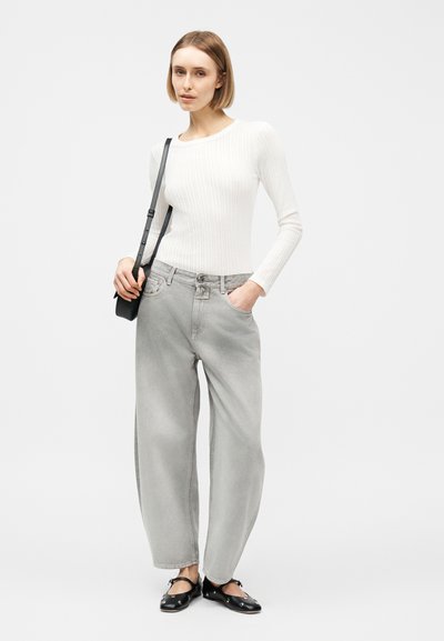 Γυναίκα με λευκό μακρυμάνικο ριμπ τοπ, χαλαρά γκρι τζιν, μαύρες μπαλαρίνες και μαύρη τσάντα χιαστί, στέκεται με το ένα χέρι στην τσέπη.