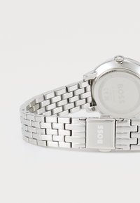 Montre en métal argenté avec un bracelet à maillons. Boîtier rond et fond, avec une couronne. Gravé "BOSS" sur le fermoir et le fond de la montre. Résistante à l'eau.