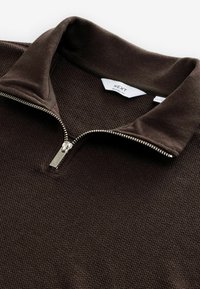 Pull en brun zippé avec une surface en tricot texturé et un col doux. Il dispose d'une fermeture éclair argentée et d'une étiquette portant la mention "NEXT Relaxed Fit."