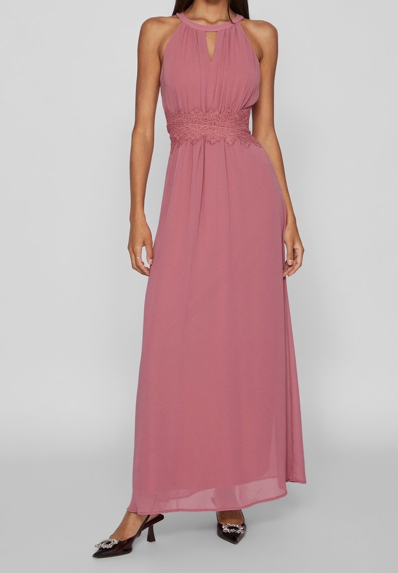 Robe maxi rose poudré sans manches avec encolure halter, taille en dentelle et jupe fluide, portée avec des escarpins noirs à bouts pointus ornés d'embellissements.