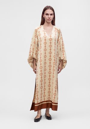 Femme portant une longue robe crème avec des motifs géométriques orange et des manches bouffantes, associée à des chaussures plates noires, debout devant un fond uni.