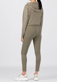 Olivgrön kort hoodie med huva, parat med figurnära olivgröna leggings och vita sportskor, visad bakifrån.