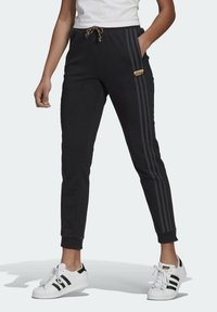 En person som bär svarta Adidas-joggingbyxor med tre mörka ränder på sidan, resårmidja med dragsko och vita Adidas-sneakers med svarta ränder.