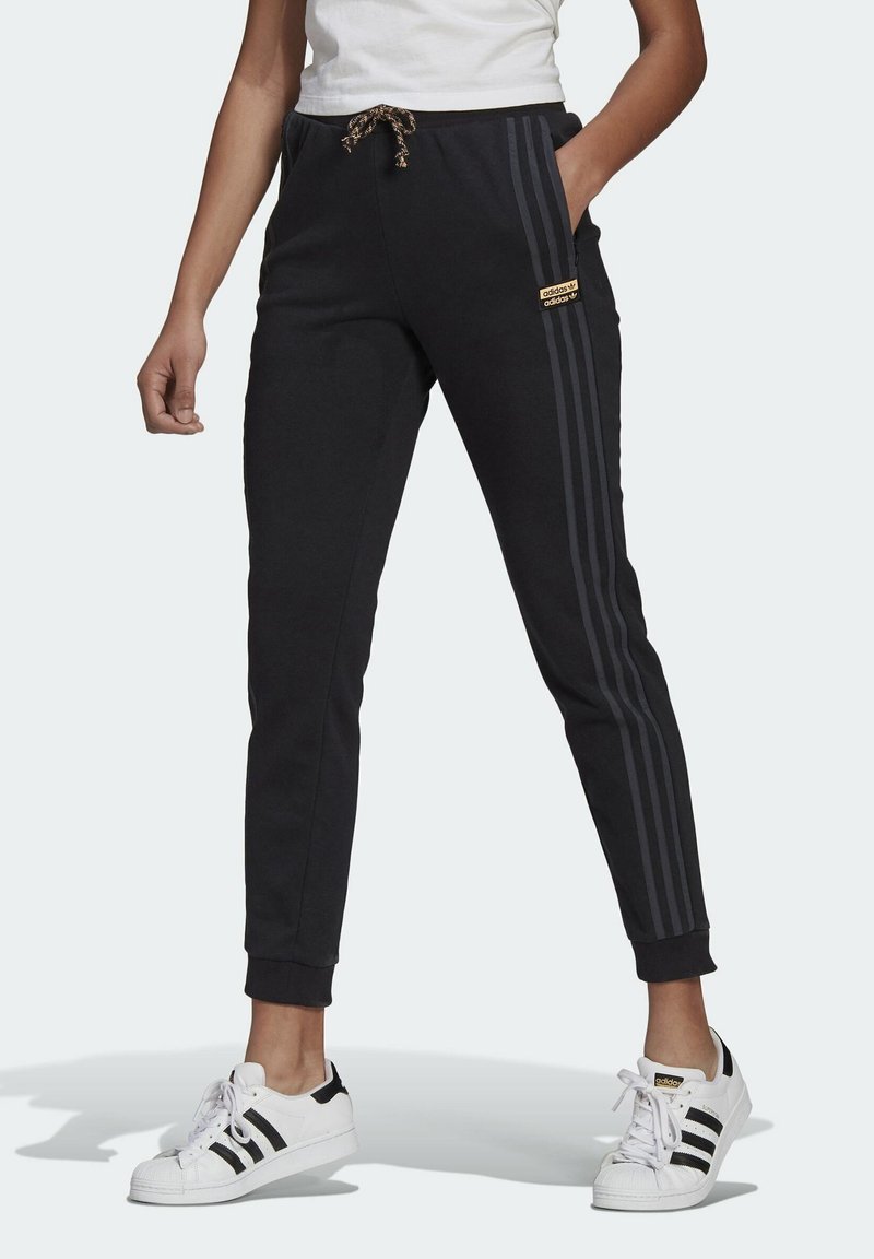 En person som bär svarta Adidas-joggingbyxor med tre mörka ränder på sidan, resårmidja med dragsko och vita Adidas-sneakers med svarta ränder.