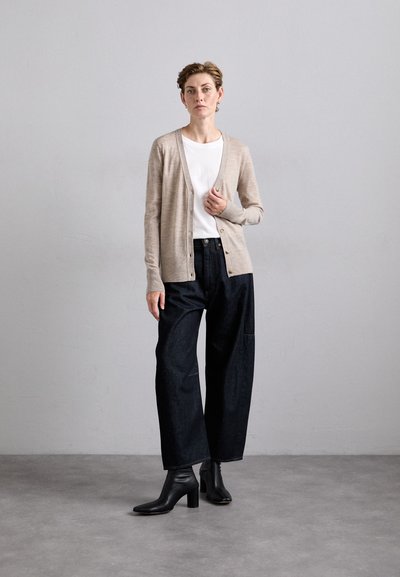 Beige gestrickte Strickjacke mit Knöpfen, getragen über einem weißen T-Shirt, kombiniert mit dunklen, lockeren Jeanshosen und schwarzen, hohen Stiefeletten.