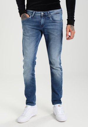 Homme portant un jean slim bleu et des baskets blanches, debout avec une main dans la poche et des tatouages visibles sur les mains.