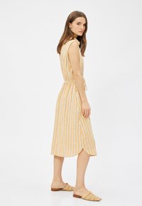 Vestido ligero sin mangas en amarillo con rayas verticales blancas. Presenta una cintura ceñida y un dobladillo a la altura de la rodilla. Combinado con zapatos de ante trenzados.