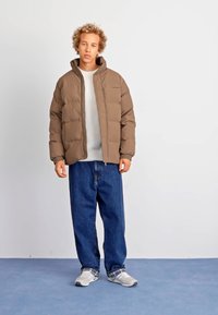 Brown puffer jacket with quilted texture, light gray sweater, loose blue denim jeans, and gray sneakers. Standing against a plain background.