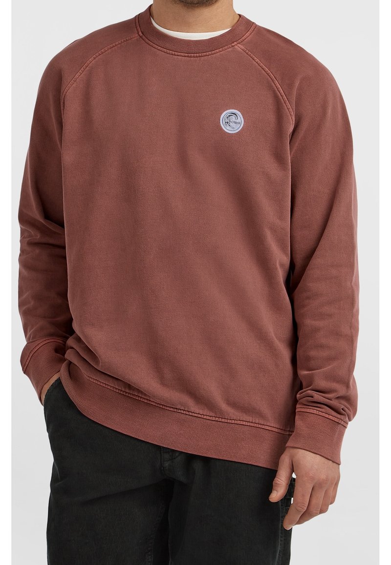 Homme portant un sweat-shirt couleur rouille avec un petit logo rond sur la poitrine gauche et un pantalon noir, main dans la poche.