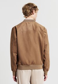 Uomo che indossa una giacca bomber in pelle scamosciata marrone e pantaloni beige chiaro, in piedi con le spalle rivolte verso la fotocamera su uno sfondo bianco.