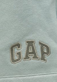 Tissu gris clair avec lettres surélevées brodées "GAP" en gris foncé, bordées de coutures gris plus clair.