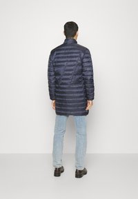 Marinblå isolerad jacka med vertikalt kviltat mönster, hög krage och långa ärmar, kombinerad med ljusblå jeans och mörka skor.