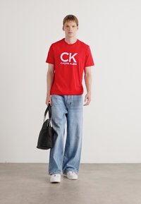 Raudona medvilninė marškinė su baltu "CK CALVIN KLEIN" logotipu, derinami su šviesiai mėlynomis plačiomis džinsinėmis kelnėmis ir baltais sportbačiais. Juoda rankinė rankoje.
