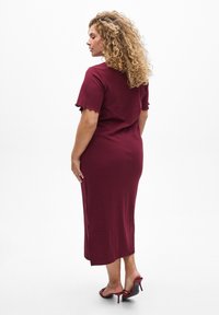 Robe midi bordeaux à côtes avec des manches courtes à bord festonné et une fente sur le côté. Portée avec des sandales à talons rouges. Texture lisse et coupe ajustée.