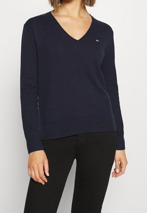 Vrouw draagt een marineblauwe V-hals trui met lange mouwen en zwarte broek, staand tegen een eenvoudige lichte achtergrond.