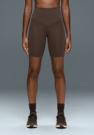 Personne portant un short cycliste marron taille haute, des chaussettes marron assorties et des baskets noires, debout sur un fond gris.