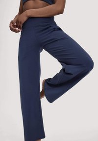 Persoon in een marineblauwe wijde broek en een bijpassend crop top, met één been iets opgetild, staand tegen een effen achtergrond.