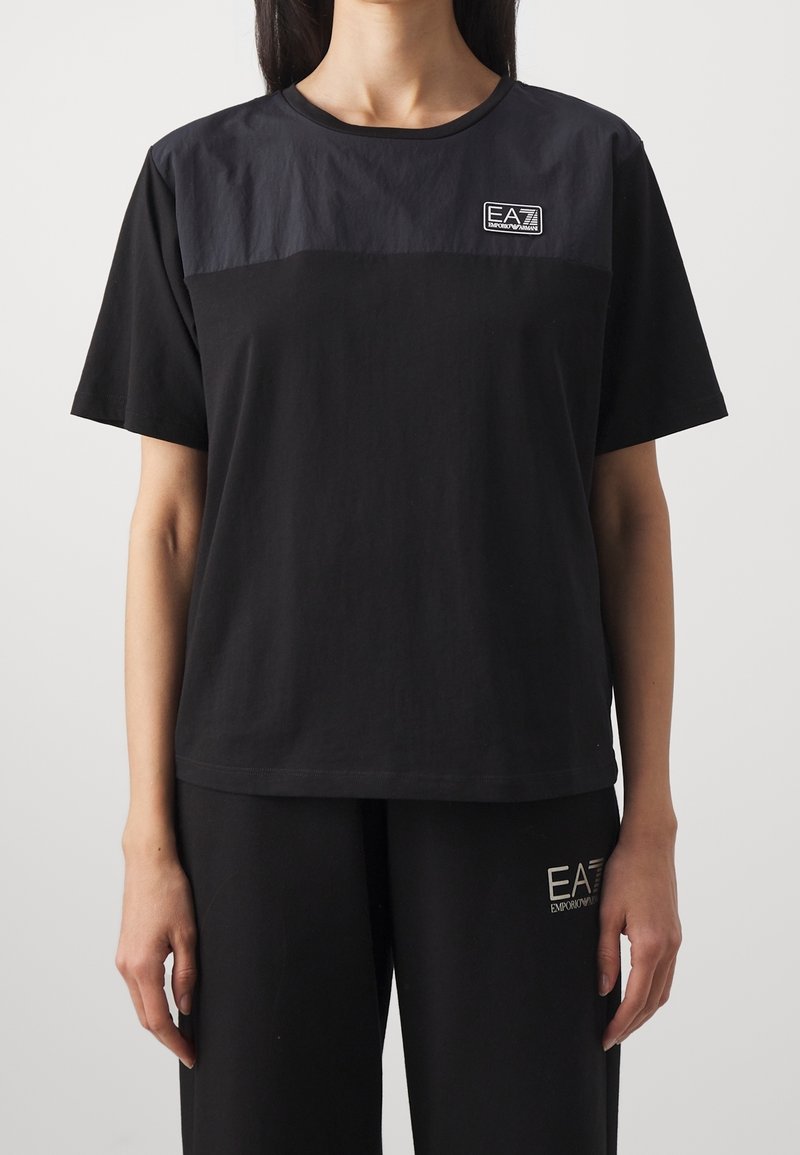 Svart bomullströja med rund hals, korta ärmar och en kontrasterande texturerad bröstpanel. Innehåller en EA7-logotyp på vänster sida.
