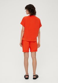 Oranje katoenmix oversized t-shirt met korte mouwen, gecombineerd met bijpassende shorts met opgerolde mouwen. Zwarte platte sandalen maken de outfit compleet.