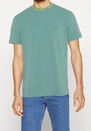 Mand iført en grøn t-shirt med korte ærmer med gul Lacoste-tekst på kraven og blå jeans, stående mod en hvid baggrund.