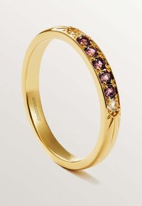Anillo de oro con banda lisa, que presenta una fila de piedras preciosas moradas y claras engastadas en un engaste de garras. Detalle de diseño acentuado en los lados.