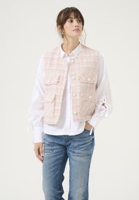 Gilet in tweed rosa con due tasche frontali e chiusura a bottoni, indossato sopra una camicia bianca, abbinato a jeans in denim blu. Design semplice e con texture.