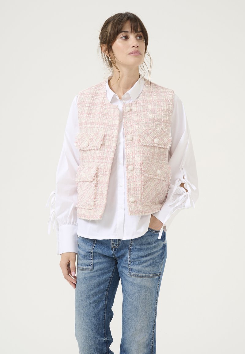 Roza tvidesta brezplačna jakna z dvema sprednjima žepoma in gumbnim zapiranjem, nošena čez belo srajco, v kombinaciji z modrimi kavbojskimi hlačami. Enostaven, teksturiran dizajn.