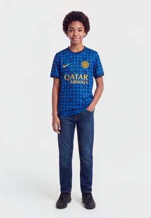 Garçon debout, une main dans la poche, portant un maillot de football bleu à motifs de l'Inter Milan, un jean foncé et des baskets noires sur fond blanc.
