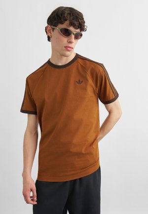 Tânăr cu păr creț purtând ochelari de soare, un tricou maro Adidas cu dungi închise la culoare și pantaloni negri, stând pe un fundal simplu.