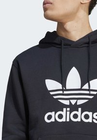 Crna dukserica s kapuljačom, sadrži bijeli trefoil logo i tekst "adidas" u podebljanom fontu. Izrađena od mekog materijala s teksturiranom površinom.