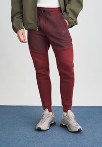 Kastanis sarkani joggeri ar kontrastējošiem tumši sarkaniem paneļiem, ar aukliņu jostasvietā, apvienoti ar pelēkiem Nike sporta apaviem. Gluda tekstūra, minimāla zīmola identifikācija.