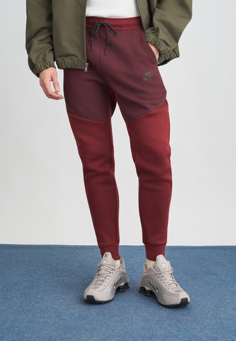 Kastanis sarkani joggeri ar kontrastējošiem tumši sarkaniem paneļiem, ar aukliņu jostasvietā, apvienoti ar pelēkiem Nike sporta apaviem. Gluda tekstūra, minimāla zīmola identifikācija.