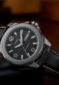 Timex analoginis laikrodis su juodu ciferblatu, sidabriniu korpusu, švytinčiais valandų žymekliais ir juoda odine apyranke su balta siūle. Atsparus vandeniui.