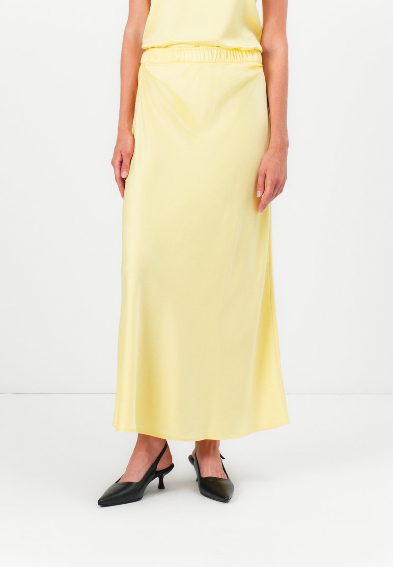 Femme portant une longue jupe en satin jaune avec une taille élastique associée à des escarpins slingback noirs à bout pointu sur fond blanc.