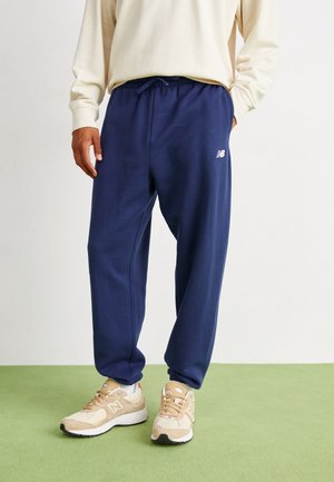 Person iført navyblå joggingbukser med elastiske manchetter og beige New Balance-sneakers, der står på grøn gulv.