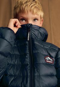 Chaqueta acolchada azul marino con un cuello alto, que presenta secciones acolchadas, un cierre con cremallera y un logo de montaña rojo y blanco en el pecho.