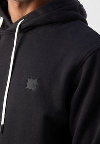 Sudadera negra de tejido suave, con un bolsillo frontal, cordones blancos y un pequeño parche de logotipo texturizado en el pecho.