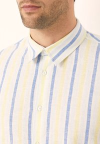 Lněná košile s modrými, žlutými a bílými vertikálními pruhy; má límeček a zapínání na knoflíky vpředu.