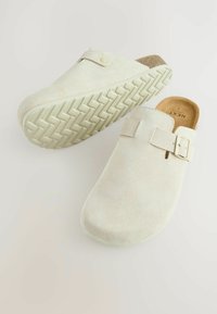Sabots en daim beige clair avec une finition lisse, une sangle réglable et une semelle en caoutchouc texturée arborant un motif en chevrons.