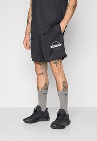 Shorts de sport en maille noire avec un logo "Celtics", associés à des chaussettes Nike grises et des chaussures de sport noires. Tatouages visibles sur les jambes.