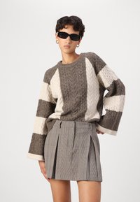 Maglione lavorato a maglia con strisce a blocchi marroni e crema e motivo a trecce, abbinato a una gonna a pieghe grigia a quadri con vita.