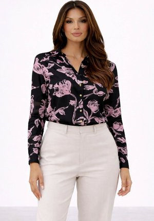 Femme portant un chemisier noir boutonné avec un imprimé floral rose rentré dans un pantalon beige taille haute, debout sur un fond blanc.
