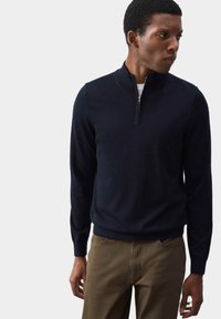 Granatowy sweter dziany z kołnierzem na pół-zamek i ściągaczami. Noszony z beżowymi spodniami, prezentującymi dopasowany krój i miękką fakturę.