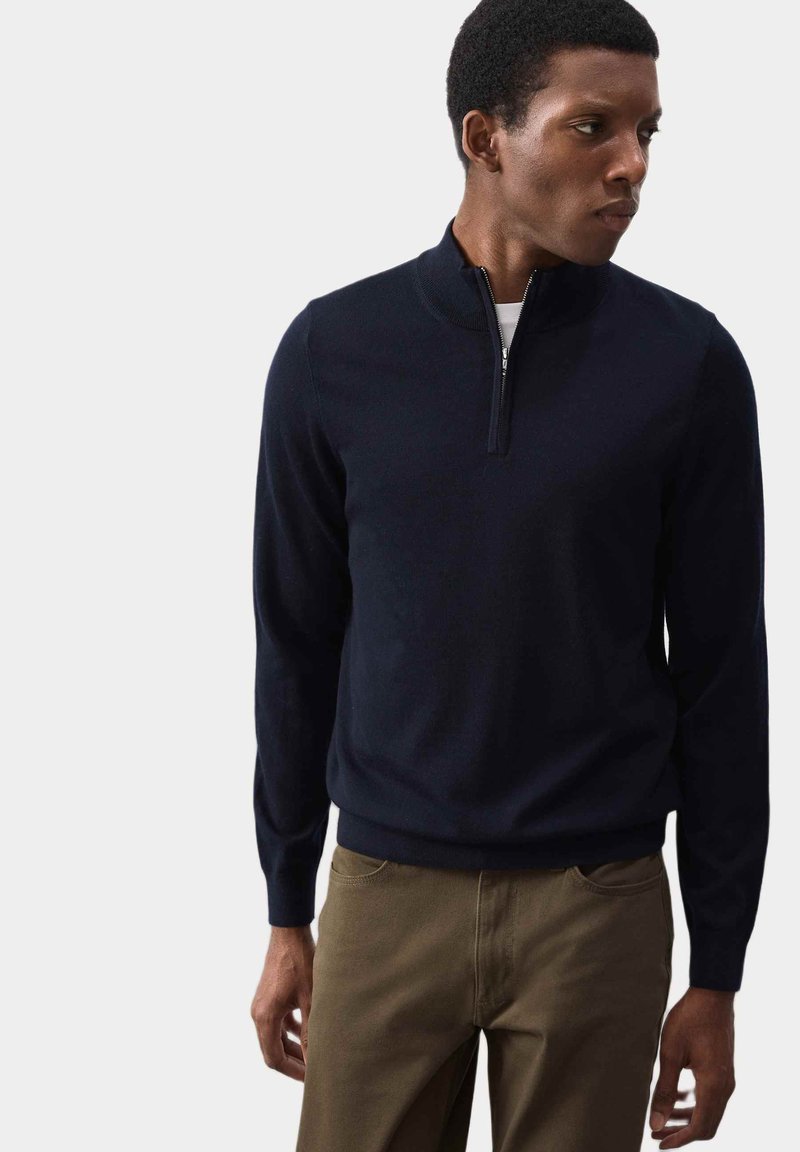 Granatowy sweter dziany z kołnierzem na pół-zamek i ściągaczami. Noszony z beżowymi spodniami, prezentującymi dopasowany krój i miękką fakturę.