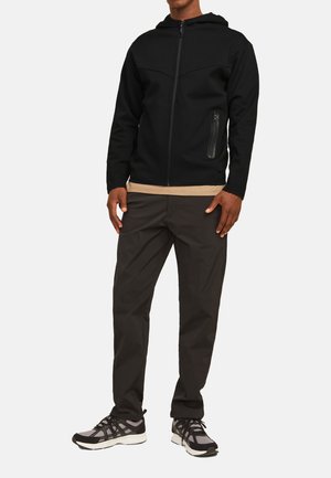 Black zip-up hoodie with a fitted design, featuring a hood and a side pocket in a textured fabric. Worn with beige undershirt and dark pants.