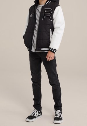 Dreng iført sort og hvid varsityjakke med stort "R", grå t-shirt, sorte jeans og sort-hvide sneakers, stående mod en ensfarvet baggrund.