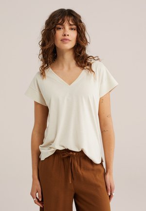 Femme aux cheveux bouclés portant un haut blanc cassé à col en V et manches courtes ainsi qu'un pantalon marron à cordon, debout devant un fond uni.